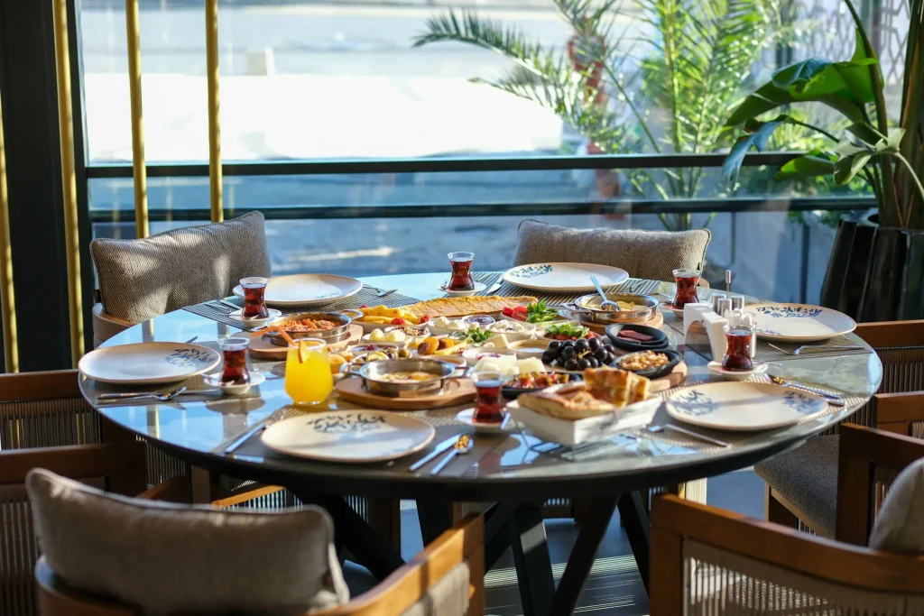 A round table is set for breakfast with bread, cheese, fruit and tea. Sunlight streams through a large window at Çakı Restoran, illuminating the table, with chairs and potted plants in the background.