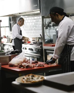 Canlı bir çakı restoran mutfağında iki şef gayretle çalışıyor: biri kırmızı bir kesme tahtasında eti doğrarken diğeri ocakta bir tencereyi karıştırıyor. Çizgili önlükler ve beyaz ceketler giymişler, şık bir çelik sıçrama tahtası ve asılı mutfak eşyalarıyla çevrililer.