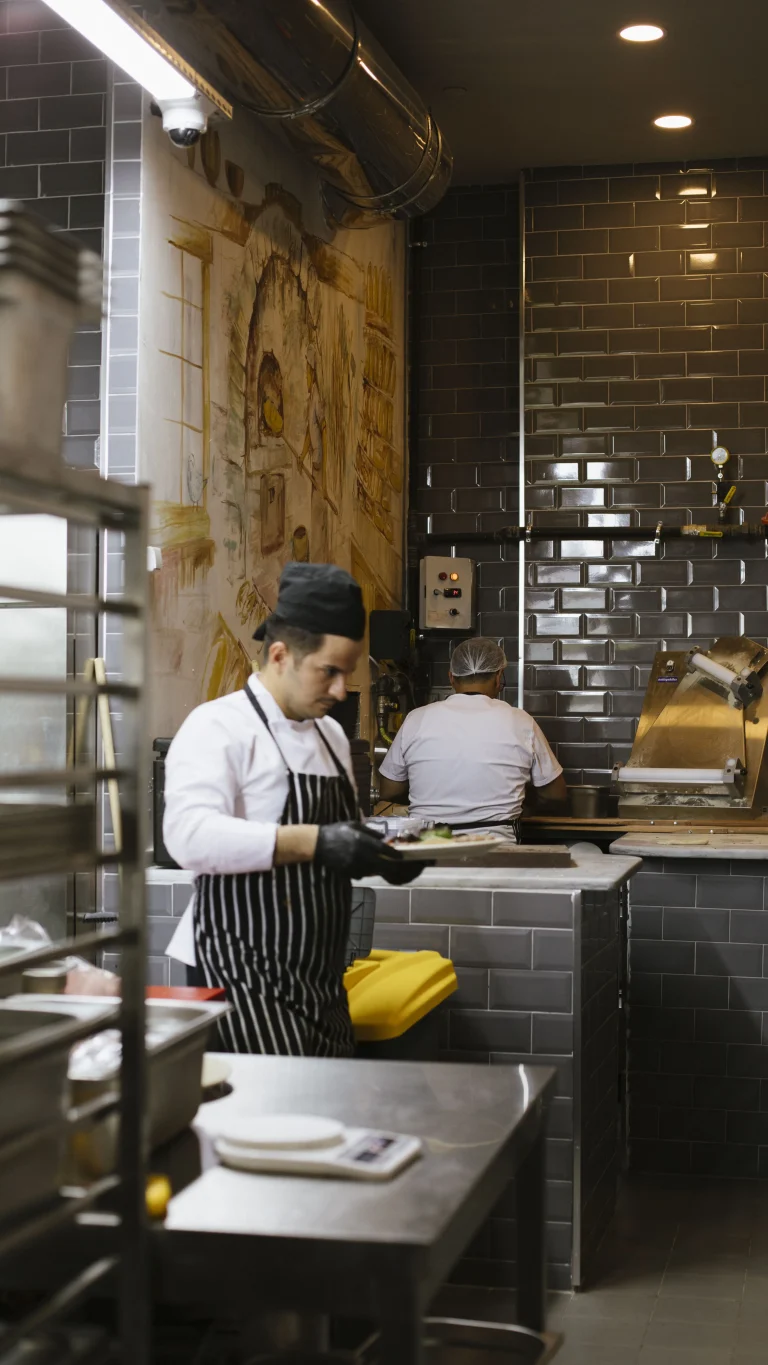 Hareketli çakı restoranında, iki şef koyu renkli fayanslı duvarlara sahip bir mutfakta gayretle çalışıyor. Ön planda siyah şapka ve çizgili önlük giyen bir şef, ustalıkla yemek hazırlıyor. Arka planda, bir diğer şef, bir dizi mutfak ekipmanının ortasında bir pizza fırınıyla ilgileniyor.