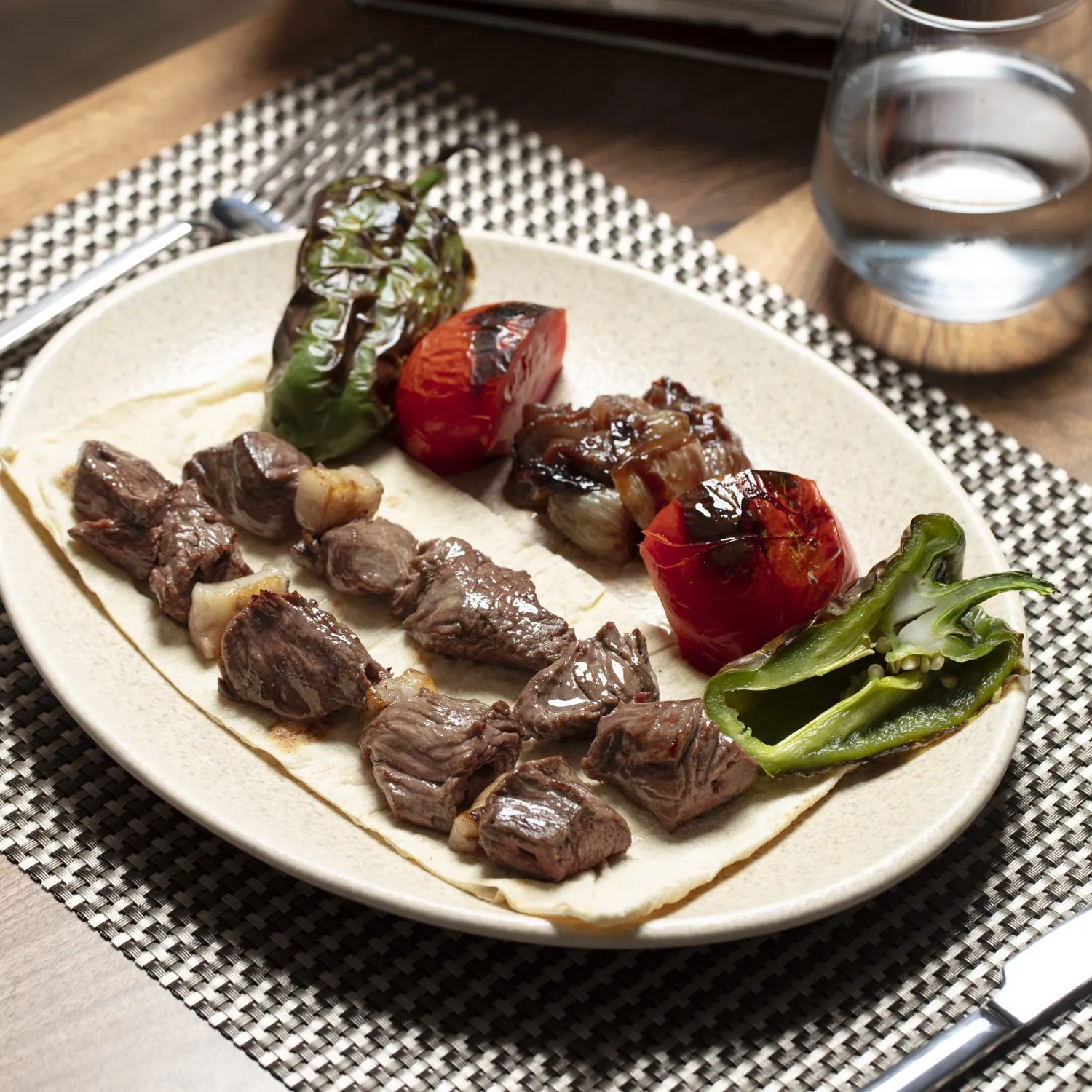 Grilled meat skewers served on flatbread with roasted green peppers, tomatoes and onions, set on a woven tablecloth with a glass of water in the background.