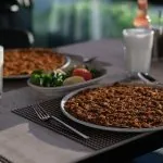 Masada içecekler, çatal bıçak takımı ve yeşillikler, limon ve domates dilimleri içeren bir salata tabağıyla birlikte iki tabak lahmacun. Arka plan yumuşak bir şekilde aydınlatılmış ve rahat bir yemek atmosferi sağlıyor.