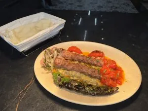 Kızarmış sebzelerin üzerinde ızgara kebaplar, kırmızı sosla tepesinde, dilimlenmiş soğan ve otlarla birlikte bir tabak. Bir sepet pide, karanlık bir tezgahın üzerinde yan tarafta.